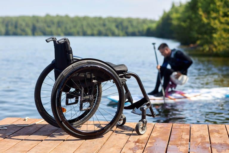 Closeup of empty wheelchair of wooden pier with man with disability enjoying water sports in background