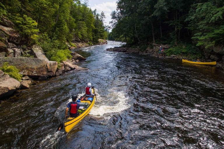 Two people wearing red life vests in a yellow canoe paddling down a river with lush greenery surrounding the river. Two people stand on the shore with another yellow canoe.